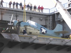 Aircraft is placed on USS Midway while people watch from flight deck.