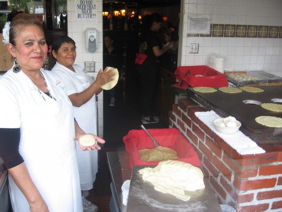 Ladies prepare fresh tortillas for people passing on the sidewalk.