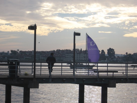 Sailboat passes fishing pier as sun falls toward horizon.
