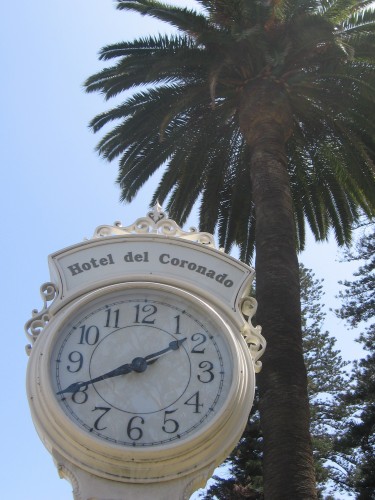 Clock stands on sidewalk among palm trees in front of hotel.