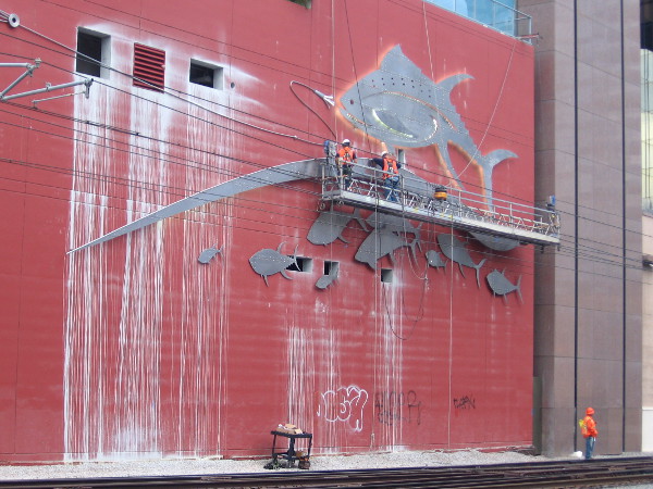 Workers on platform busy mounting a gigantic trophy fish to a high-rise building!