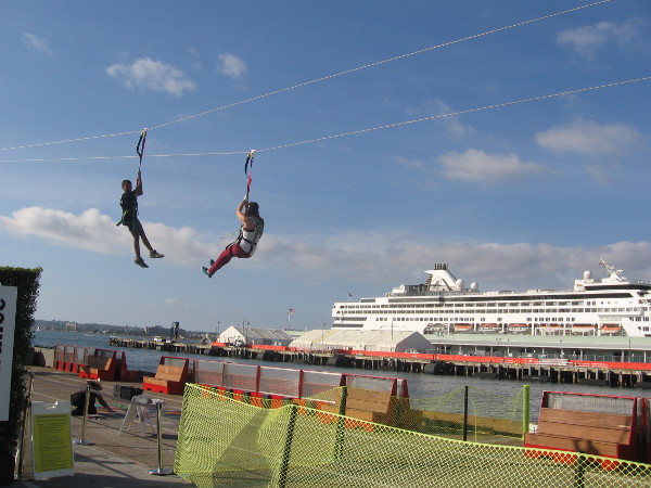 Brave souls soar down a zip-line with a docked cruise ship in background.