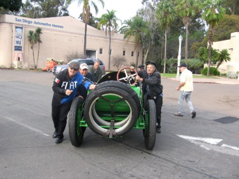 Old race car is pushed into position. San Diego Automotive Museum is in the background.