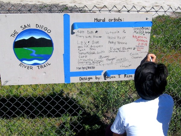 Mural artists sign their names on sign beside the San Diego River Trail.