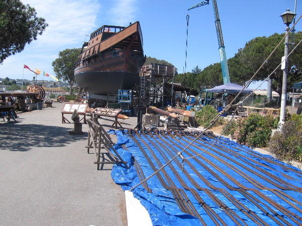Suspended from a crane is a shroud (rigging that helps hold a mast) that's being tarred. The bow of the galleon comes to a sharp point at its beak.
