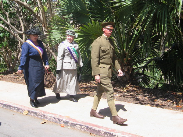 Two suffragettes with Votes For Women sashes follow a guy dressed in World War I era military uniform.