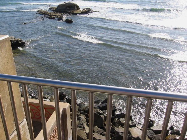 Looking down over the view point rail at rugged rocks at the edge of the shining Pacific Ocean.