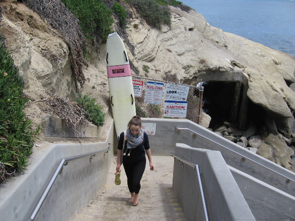 A lady climbs stairs up from the beach, past a lifeguard's rescue board.