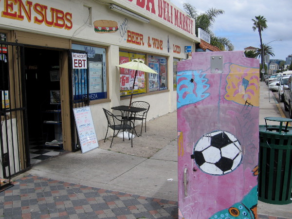 A soccer ball on utility box by a small local market known for their sub sandwiches.