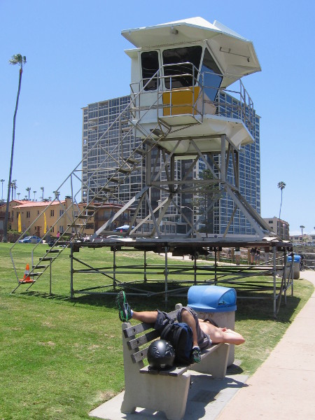 Guy takes a nap on bench beneath a lifeguard tower.
