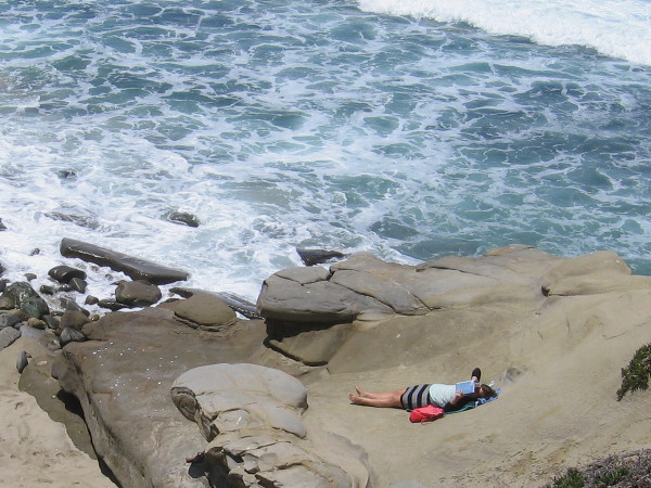 A lady lies on a flat rock reading a book, as waves crash nearby.