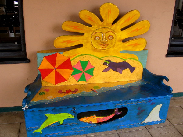 A happy sun shines above beach and ocean on this unique bench, where anyone might sit and relax.
