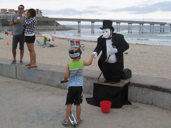 A masked street performer shakes hands with a kid on a scooter. The Ocean Beach Municipal Pier stretches in the background.