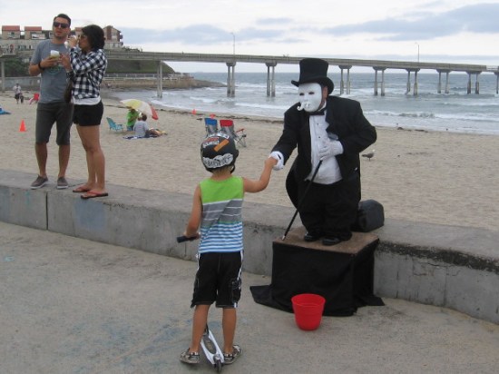 A masked street performer shakes hands with a kid on a scooter. The Ocean Beach Municipal Pier stretches in the background.