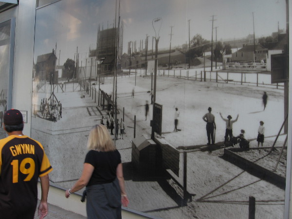 Padres fans walk down 11th Avenue toward Petco Park on a game day. They pass a mural featuring photos of baseball in San Diego many years ago.