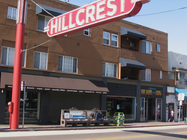 An artistic trashcan waits by a bus stop near the Hillcrest landmark sign.