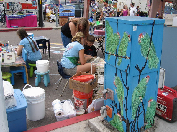 Young artists use their imaginations as they work behind a utility box decorated with parrots in Ocean Beach.