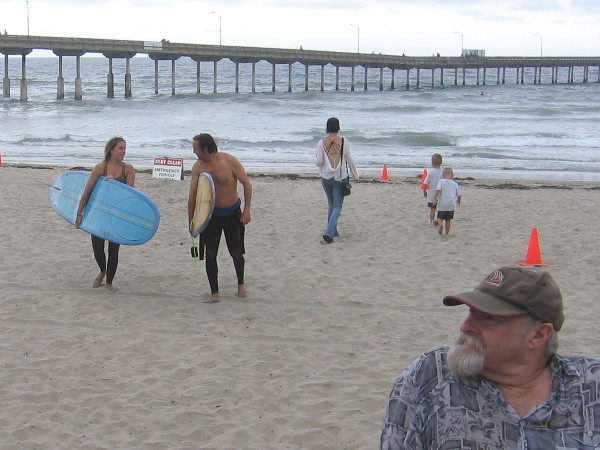 Surfers come up across the beach while a guy relaxes on the sea wall.
