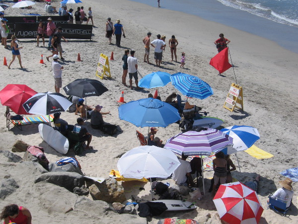Folks in the shade of beach umbrellas look toward the surf, where dueling Supergirls are performing astonishing feats during 25 minute heats.