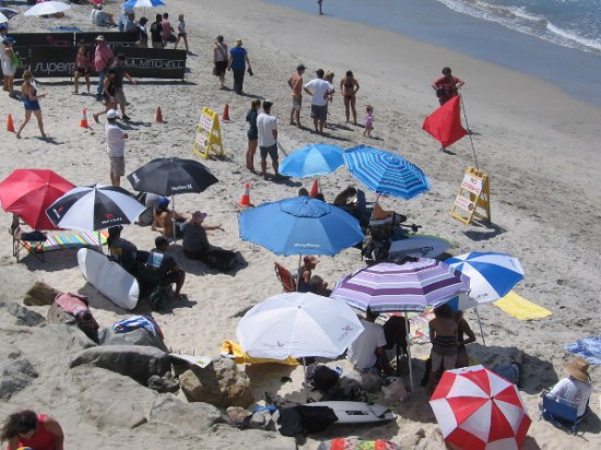 Folks in the shade of beach umbrellas look toward the surf, where dueling Supergirls are performing astonishing feats during 25 minute heats.