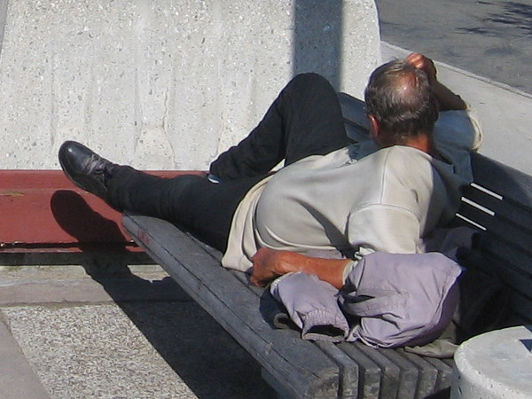 Homeless man stretches out on a public bench beside the water.