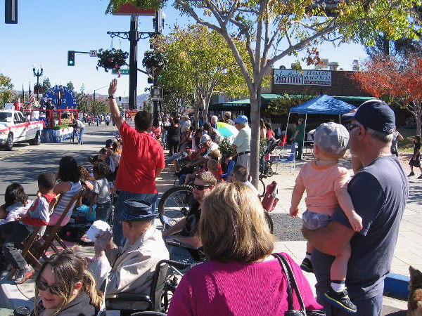 Families have gathered on a beautiful Saturday morning. Kids and adults watch the Mother Goose Parade, which this year has a super hero theme.