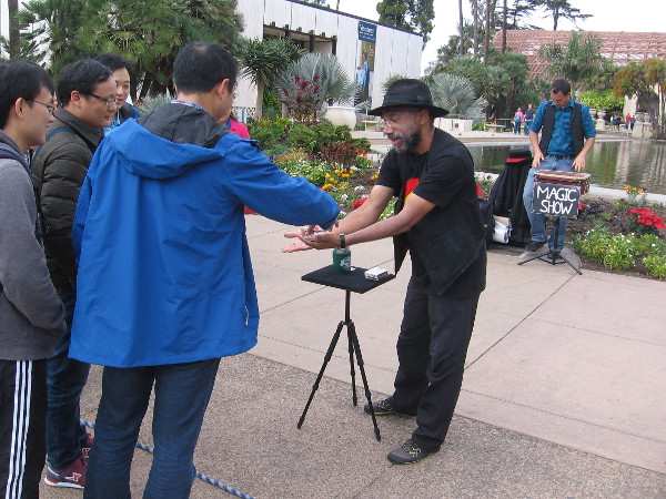Tourists are shown magic by a fun busker near the reflecting pond in Balboa Park.