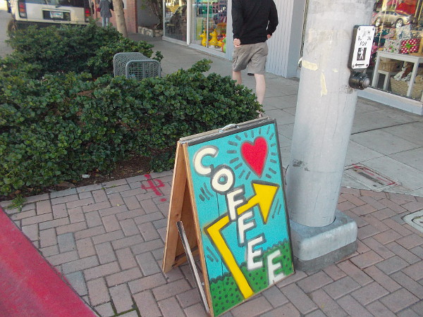 A bold, artistic sign entices people to grab a cup of coffee nearby.