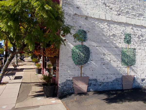 Greenery painted on an old brick wall seems to blend with lush trees along the sidewalk.
