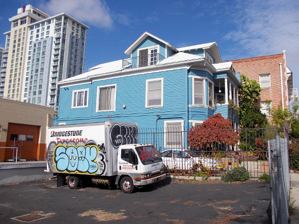 Graffiti on a truck, an old blue house, and a modern high-rise, together in one photo.