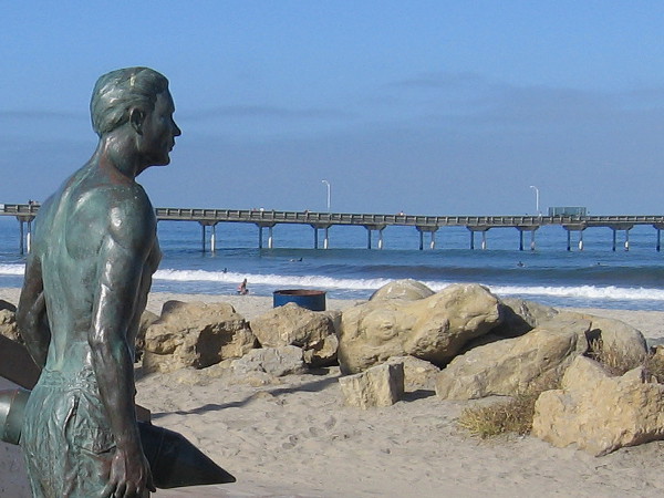 A bronze hero stands guard on the sand in Ocean Beach. The long OB pier stretches out into the Pacific Ocean in the background of this photo.