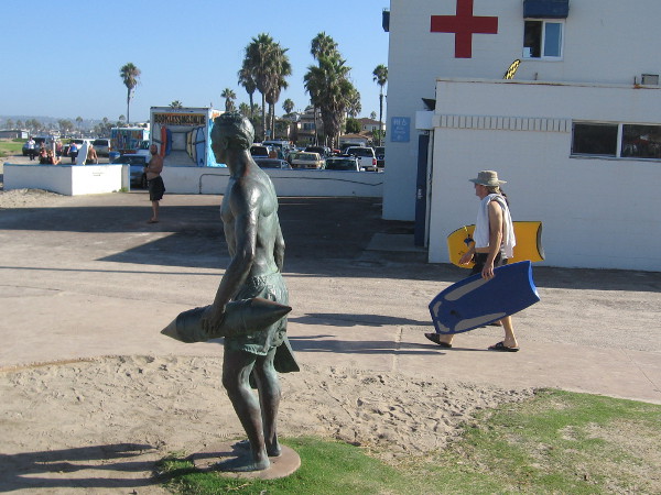 Beachgoer with two boogie boards passes between the bronze sculpture and the main Ocean Beach lifeguard station.