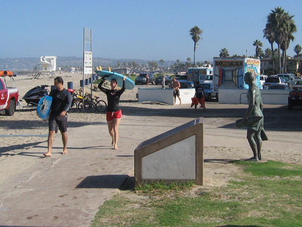 A couple of surfer dudes carrying surfboards approach the memorial plaque, a few feet from the lifeguard tribute statue.