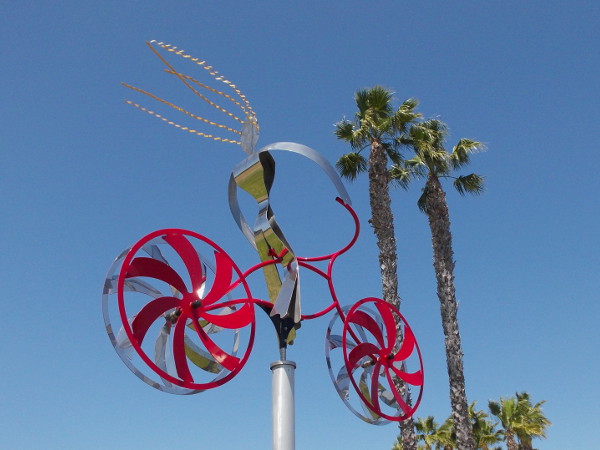 A steel bicyclist with wildly blowing hair flies through the blue sky past some Southern California palm trees!