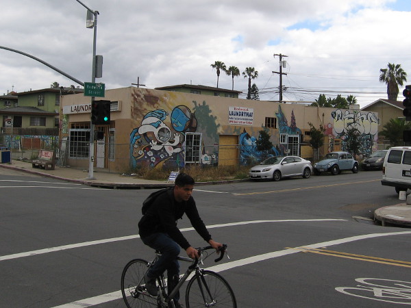 Bicyclist rides south down 30th Street in North Park, past an old closed laundromat at Redwood Street. This building's walls have long featured diverse and changing works of street art.