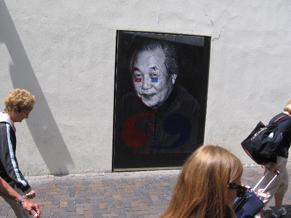 People walk through the breezeway between MCASD and the Santa Fe Depot. Three large portraits address the theme of political and domestic violence.