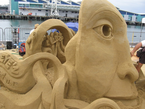 Small people stand together behind a mask-like face made of sand.