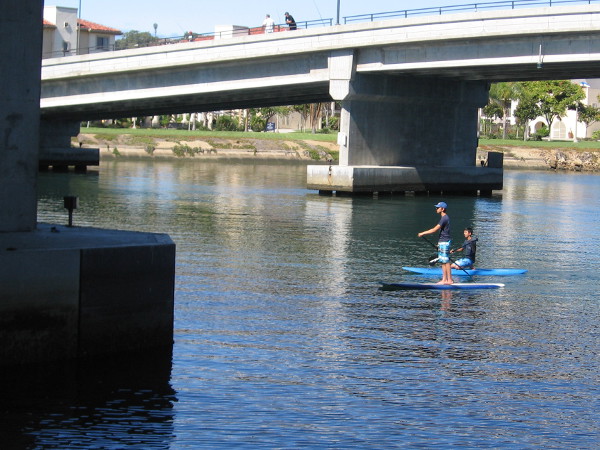 Paddleboarders float down the boat channel between North Harbor Drive Bridge and the adjacent Nimitz Bridge, which is now used by pedestrians. The grass in the distance is part of the Liberty Station Esplanade.
