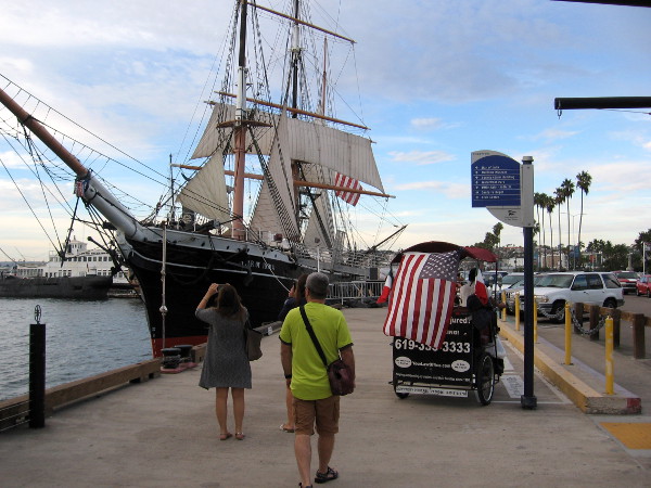 Pedestrians and a flag-draped pedicab approach the famous tall ship Star of India at the Maritime Museum of San Diego.