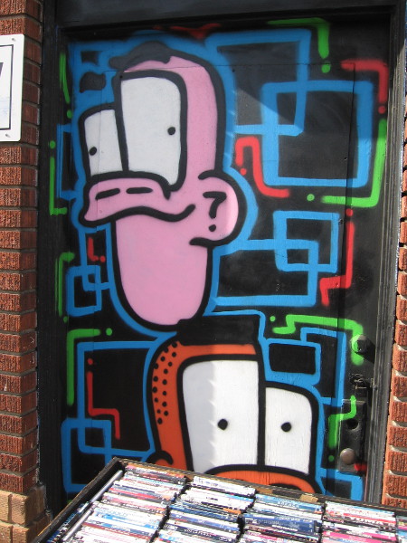 Two funky faces watch people walking down the sidewalk from a window above a bin of CD's.