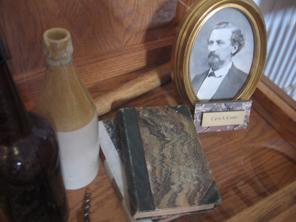Historical artifacts in a glass display case include books, bottles and a photo of Cave J. Couts.