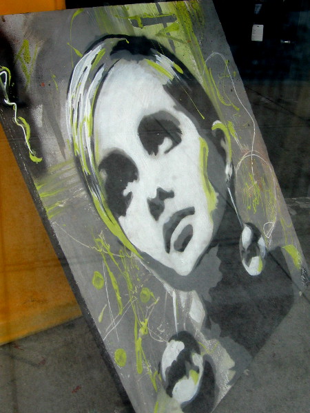 Painting of female face in window of a small shop in East Village containing odd bits of art and miscellaneous used items.