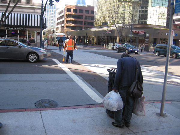 A worker crosses Broadway while a homeless man looks into a trashcan.