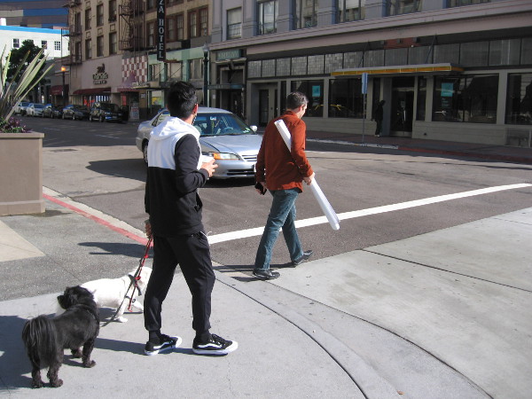 One guy crosses the street while carrying plans, another escorts dogs with a coffee in hand.