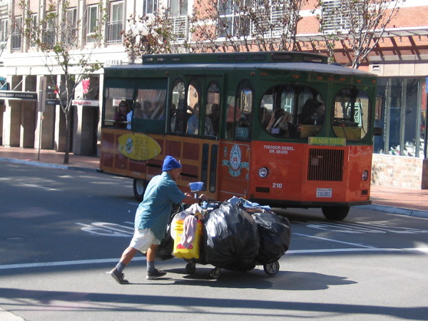 An Old Town Trolley Tours vehicle loaded with tourists waits for a homeless man with a packed shopping cart to clear a downtown intersection.