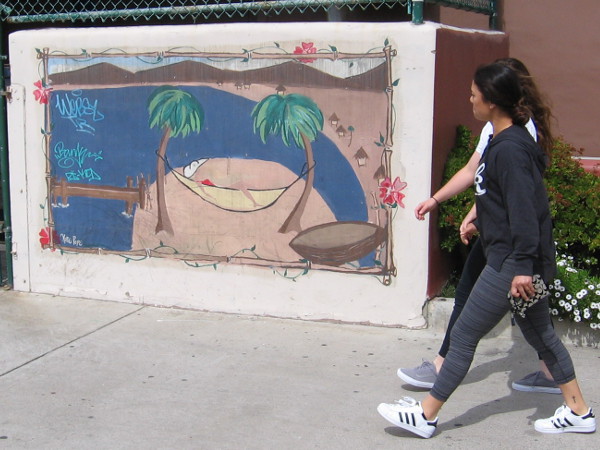 Two ladies walking along the edge of a parking lot toward the nearby beach. They pass someone snoozing in a hammock strung between palm trees.