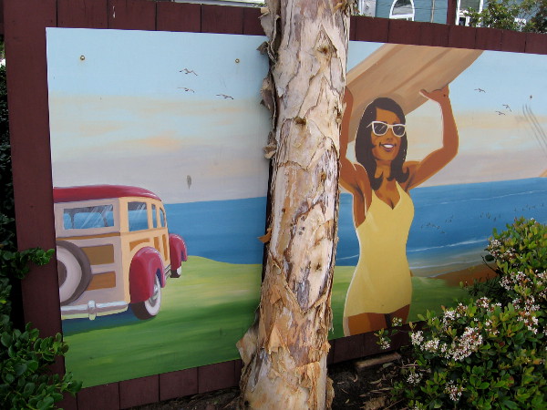 One of several cool, nostalgic beach images on one parking lot fence. A surfer girl and a woodie overlooking the ocean.