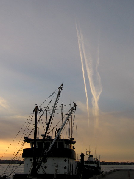 Aircraft condensation trails catch the sun high in the sky, as light fades around fishing vessels docked at the G Street Pier.
