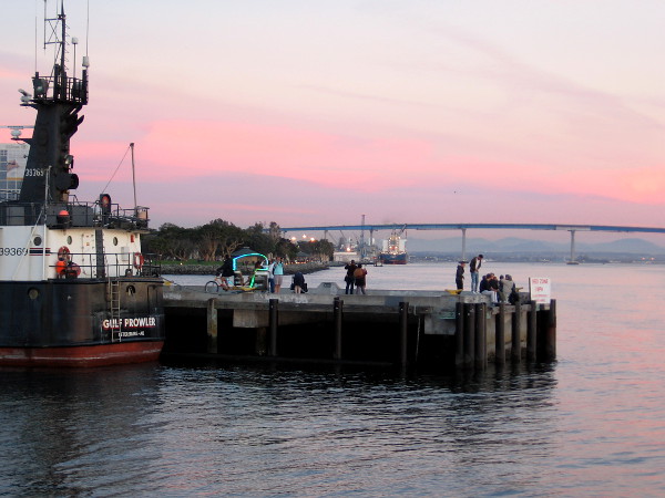 People linger into the evening at the end of the pier near Seaport Village. A pedicab with colorful lights turns around near a docked ship.