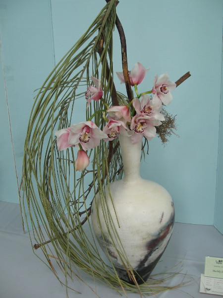 A fine vase containing orchids, palm leaves and dry branch.